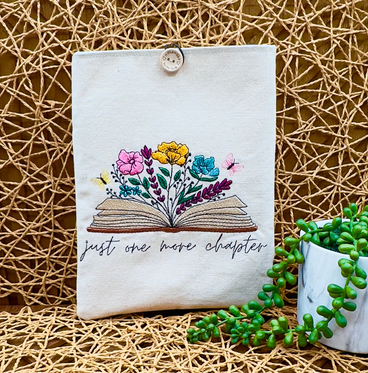 light beige canvas book sleeve with a flowering book embroidered along with "just one more chapter" embroidered below the book, features a wood button closure