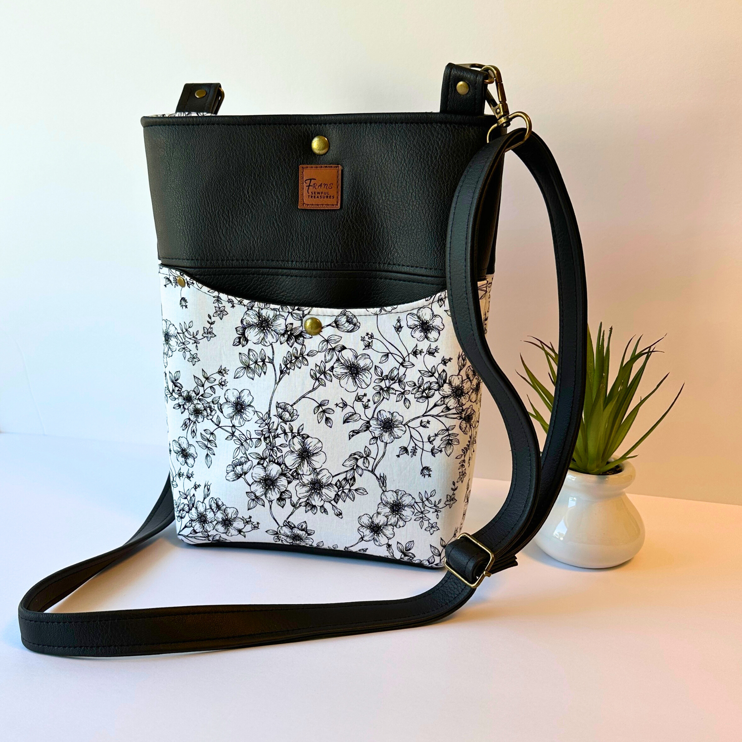 black vinyl  featuring white with black floral print crossbody bag with a detachable and adjustable strap.  Antique brass hardware
